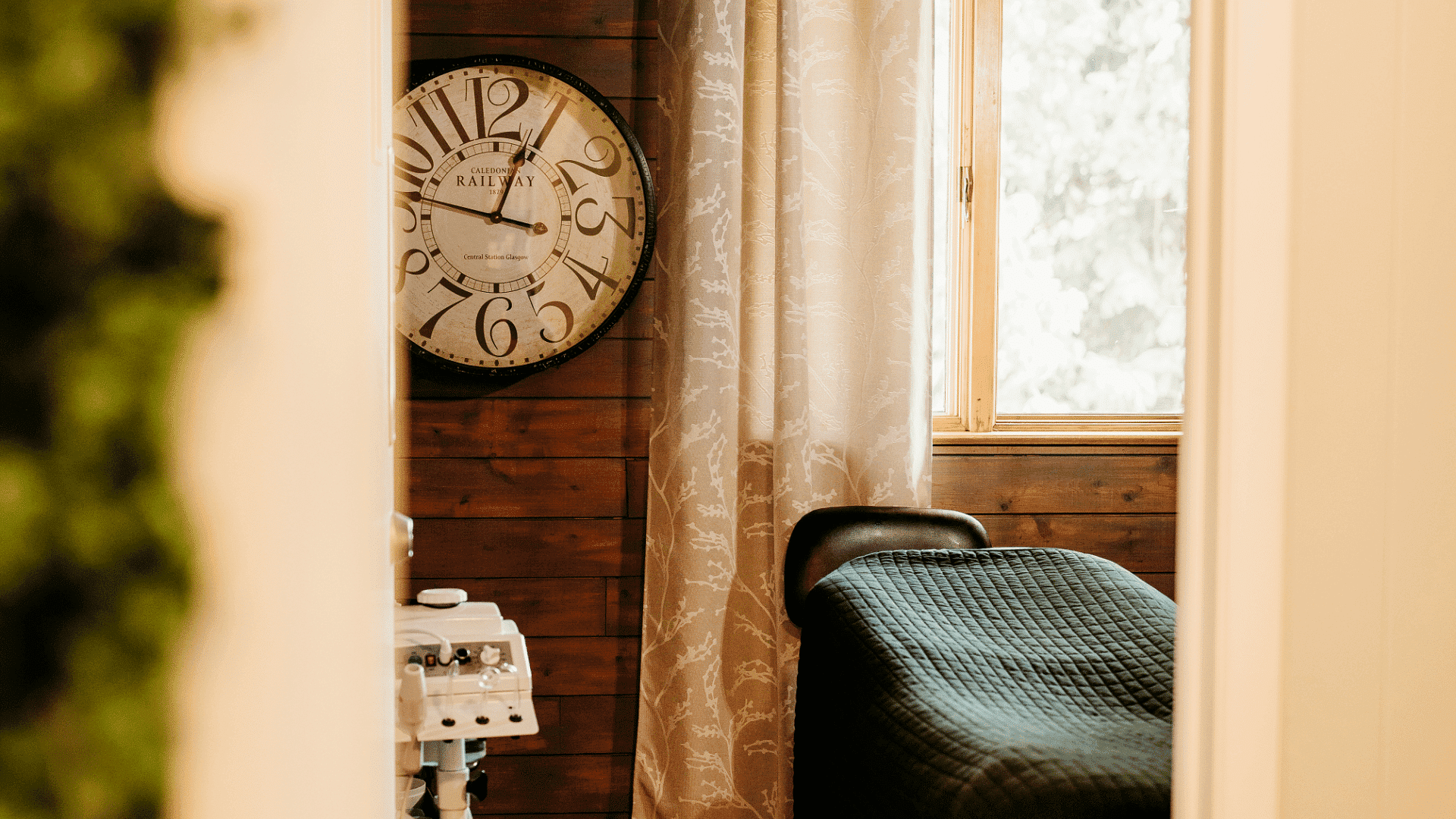 Cozy spa room with a massage table, vintage clock, and a window with sheer curtains.