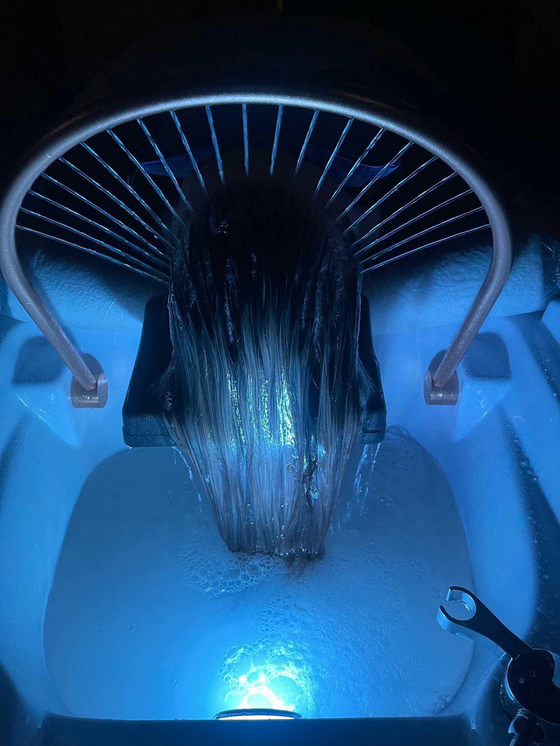 Person's head under water stream in a futuristic hair washing station with blue lighting.