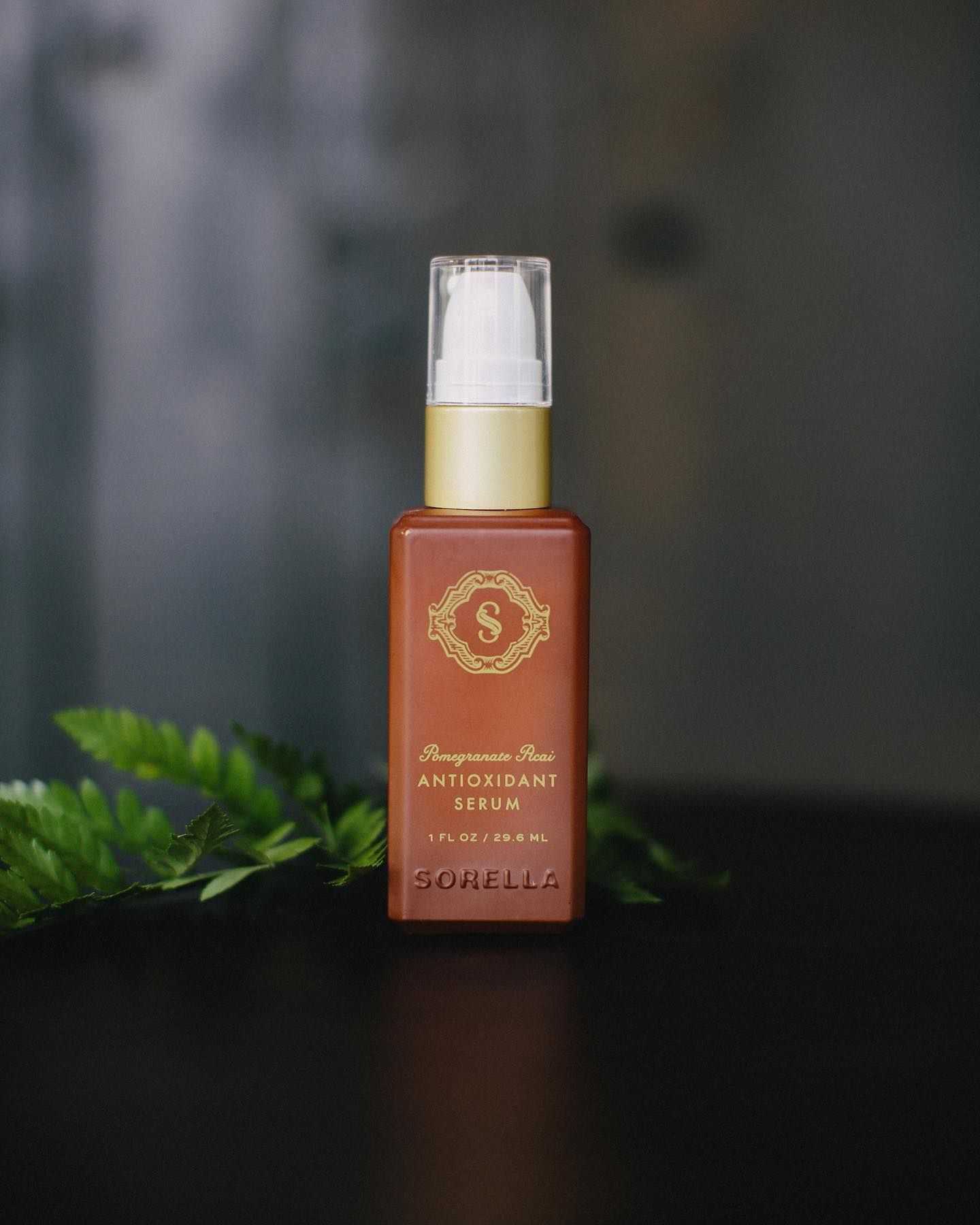 Red bottle of Sorella Pomegranate Acai Antioxidant Serum on a black surface with green leaves.