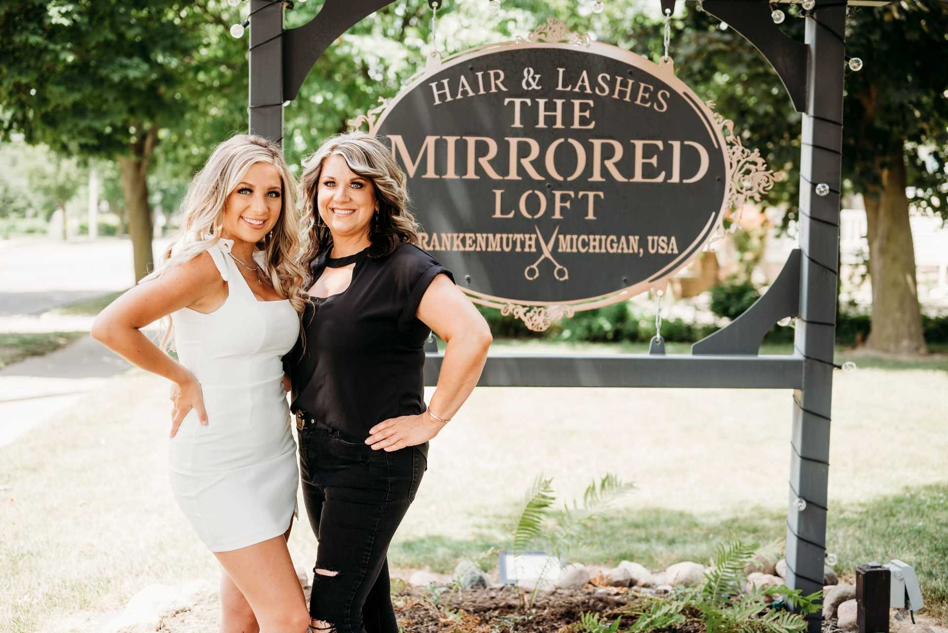 Two women smiling in front of "The Mirrored Loft" hair and lashes salon sign in Frankenmuth, USA.