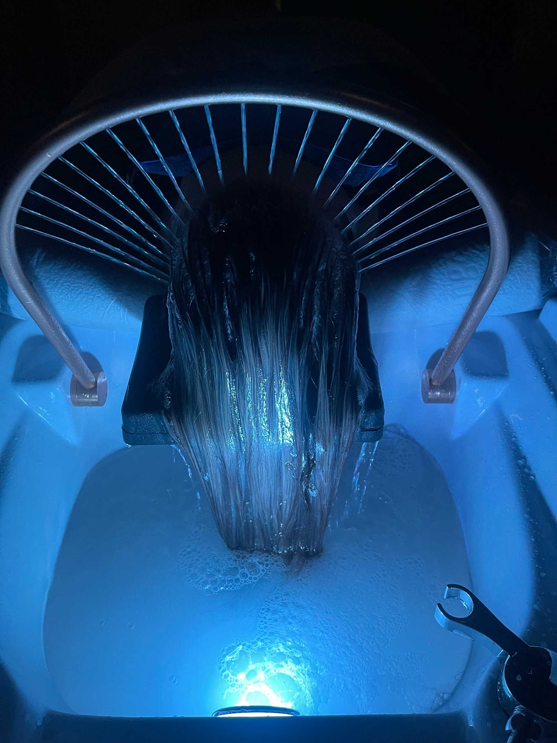 Person's head under water stream in a futuristic hair washing station with blue lighting.